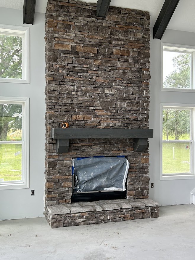 Stone fireplace with wood mantel, grey tarp covering adjacent window, stone wall detail, window framing view of trees outside