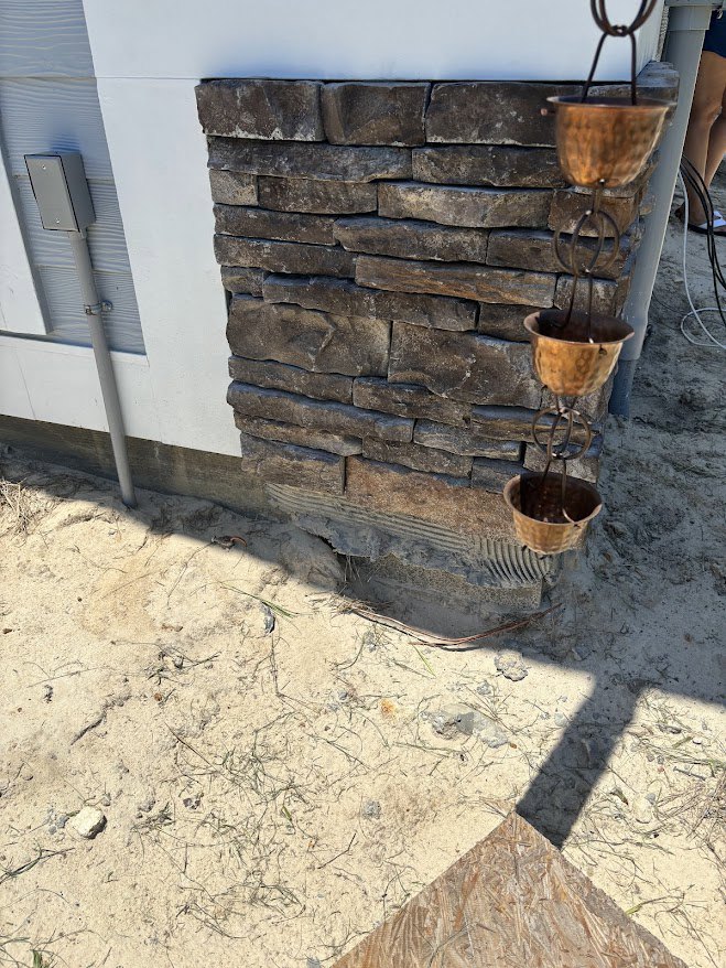 Rain chain with metal bowls hanging from a brick exterior wall, white downspout pipe visible, stone ground below