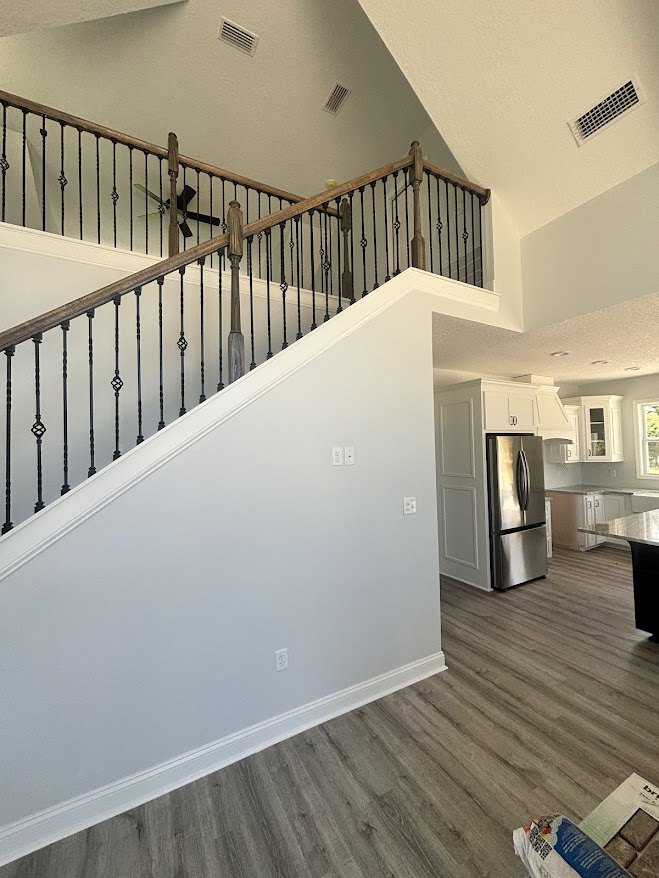 Wood and metal staircase with black handrail, light plaster walls, hardwood flooring, and stainless steel refrigerator in adjacent kitchen area
