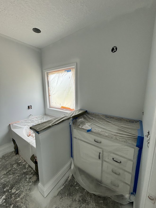 Bathroom with freestanding tub and sink, white dresser wrapped in plastic, close-up of smooth white plaster wall, light flooring, cabinetry with drawers.