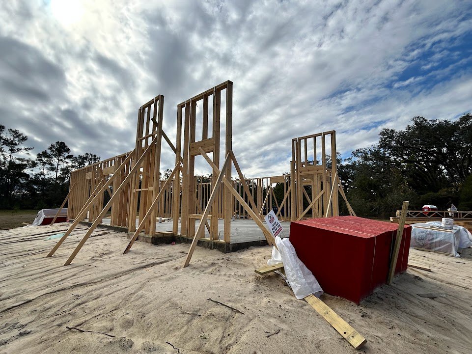 Wood-framed house under construction with exposed beams, stacks of lumber, red toolbox on sandy ground, white bag on wooden board, cloudy sky overhead