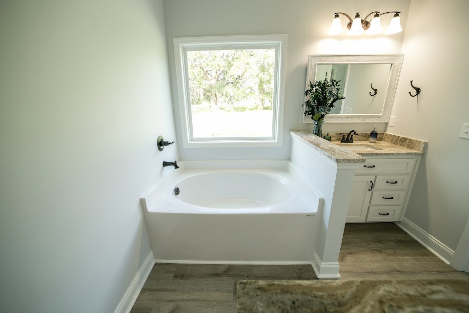 White freestanding bathtub with matte black faucets, vessel sink on wood vanity, large mirror reflecting vase of flowers, three-light fixture above, window with view of trees