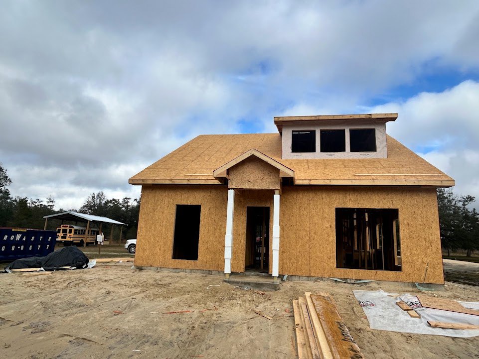 Wood-framed house under construction with exposed beams and plywood, surrounded by dirt and construction materials, set against a cloudy sky