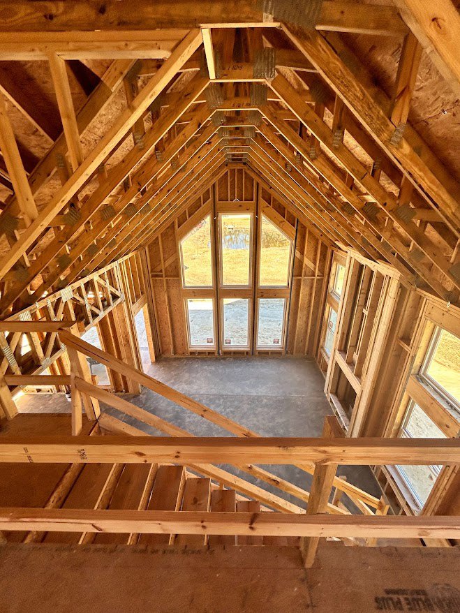 Exposed wooden beams and framing inside a house under construction, with a staircase, window, and brown wall featuring a wooden railing