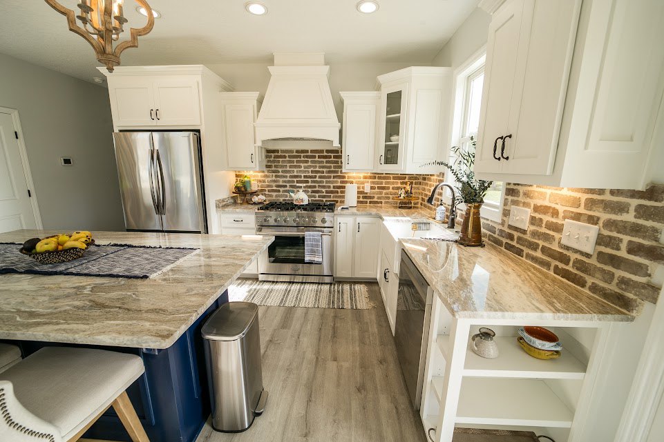 Marble kitchen island with built-in sink and faucet, exposed brick wall backdrop, stainless steel refrigerator, metal container on countertop, wooden chair with white cushion