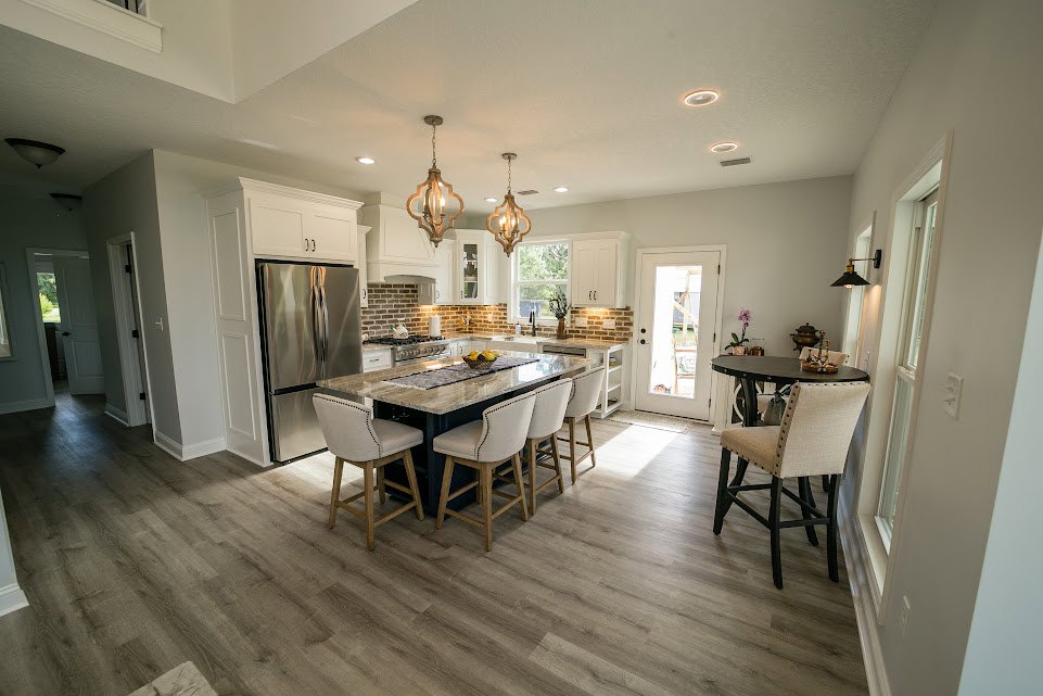Open kitchen with a large central island featuring barstools and a basket of fruit, adjacent dining table with chairs, stainless steel refrigerator, light wood flooring, white