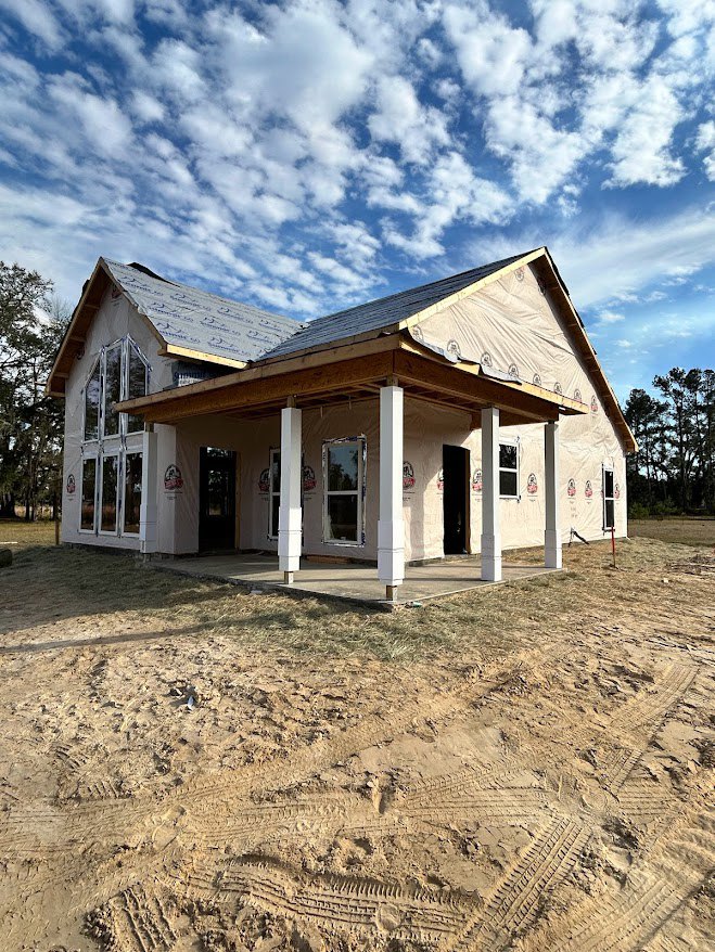 Wood-framed house under construction with covered porch, tire tracks in sandy dirt foreground, group of trees in background, roof beneath partly cloudy blue sky