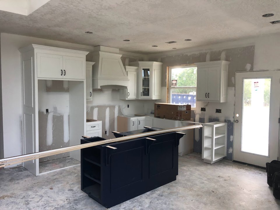 White kitchen cabinets, black island with wood trim, lighted ceiling, windowed door, empty white shelving, box-shaped pendant light over island.