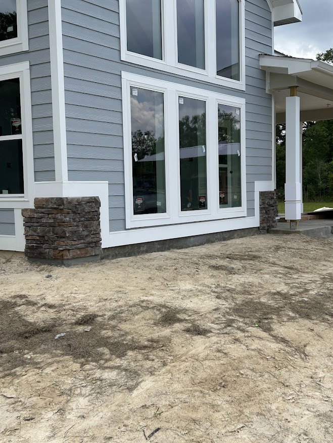 Stone exterior wall with sash windows, white pillar on porch, dirt ground surrounding unfinished custom home
