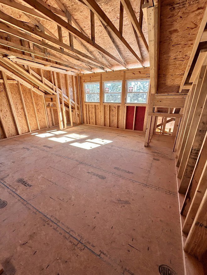 Exposed wood ceiling beams, large windows, unfinished wood floor, and visible framing in a residential interior under construction