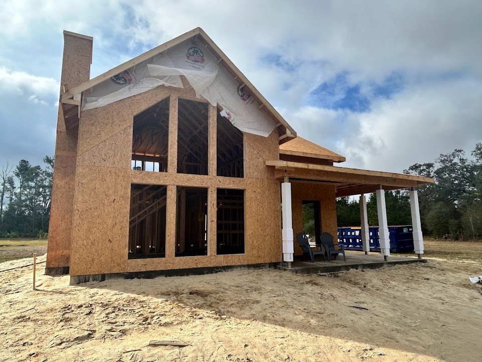 Partially built house with exposed lumber framing, chimney, dirt yard, scattered chairs, blue container marked with white numbers, and clear blue sky overhead