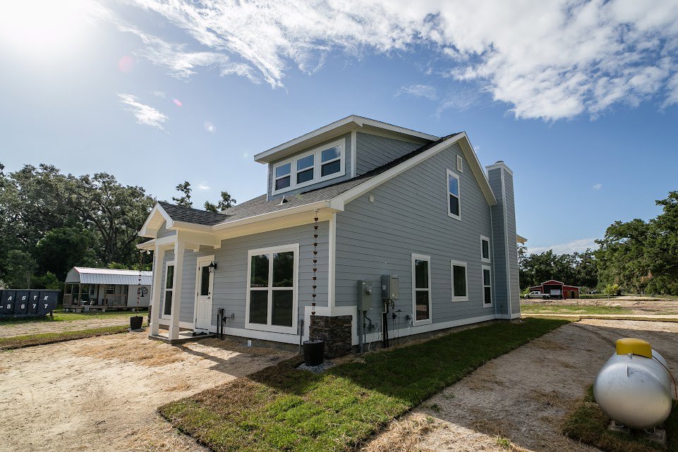 Single-story home with white-framed windows, covered porch, wind chime, large silver tank with yellow top, dirt path leading to entrance, surrounded by trees under a partly cloudy