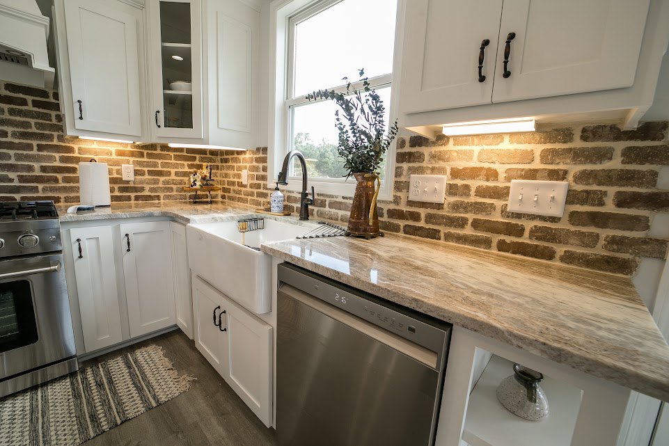 Brick-walled kitchen with marble countertop, stainless steel dishwasher, copper pitcher holding a plant, close-up microwave, four-switch white light switch, and decorative vase