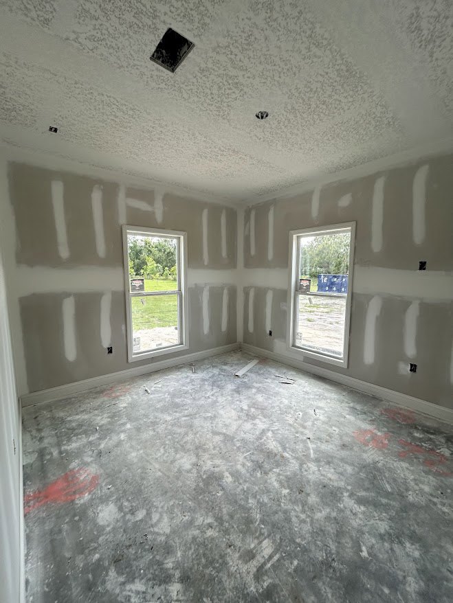White-walled room with large windows, concrete floor, black rectangular object on a white surface, view of grassy field through window