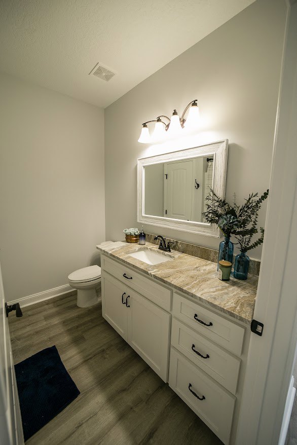 Bathroom with marble countertop, large mirror, white toilet, blue vases holding green plants, light tile flooring, and wood cabinetry with drawers.