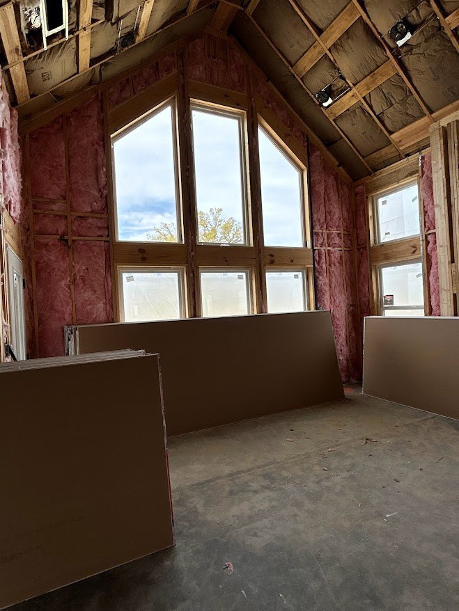 Room with exposed wood framing, large windows overlooking trees, concrete floor with scattered dirt, and a person standing near a brown wall.