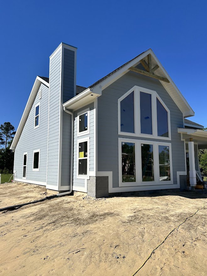 Framed house under construction with exposed wooden beams, unfinished siding, dirt ground, and clear blue sky; close-up of window installation visible
