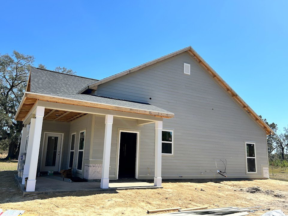 Partially built house with white siding, black framed doorway, covered porch, dirt yard, and dark shingle roof under clear blue sky