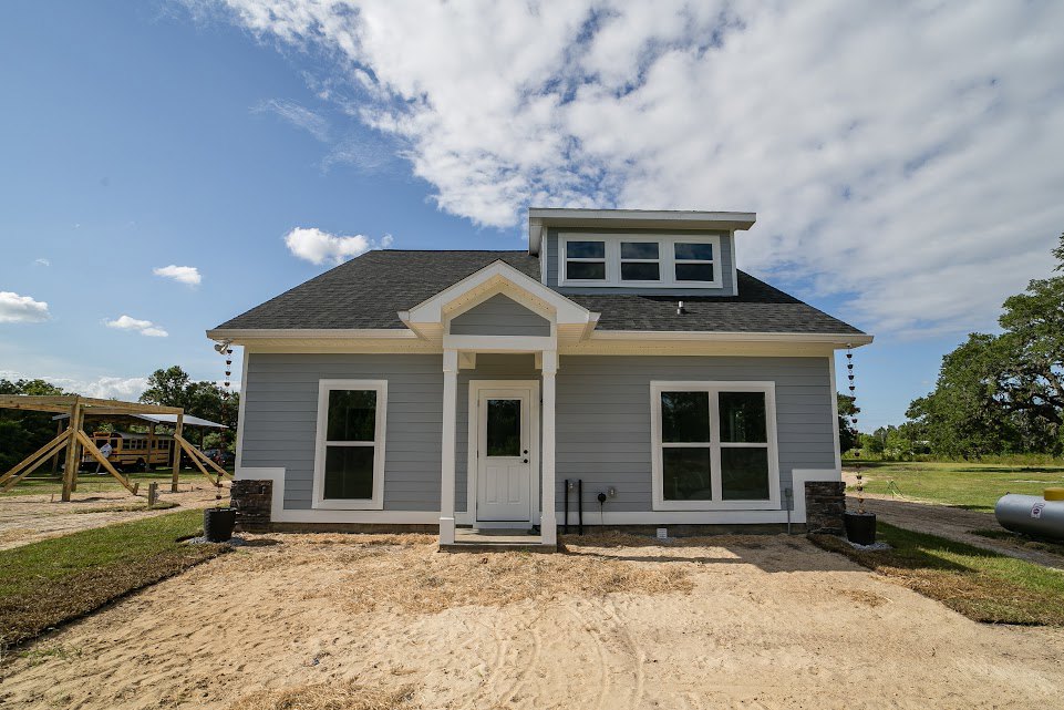 Partially built house with exposed framing, white door with window, large windows with white frames, sandy ground with tire tracks, surrounding trees and cloudy sky in background