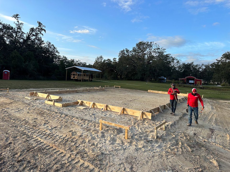 Two men standing on sandy ground beside a partially constructed building with exposed wooden framing, a bus parked under a roof, and scattered construction materials.