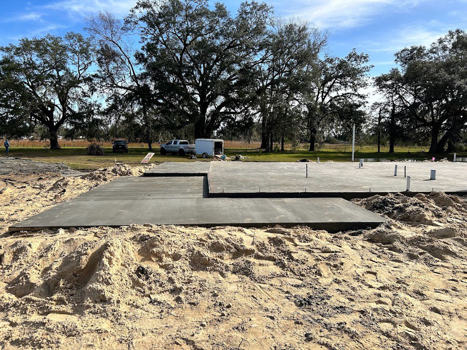 Concrete slab set in sandy soil near a large tree with sprawling branches, blue sky and scattered clouds overhead, parked truck under tree, surrounding group of trees.