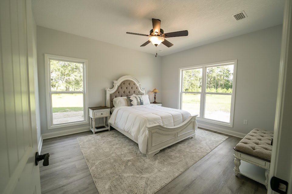 Bedroom with a white bed frame, ceiling fan with light, bench at the foot of the bed, window showing trees outside, and white table with black drawer.