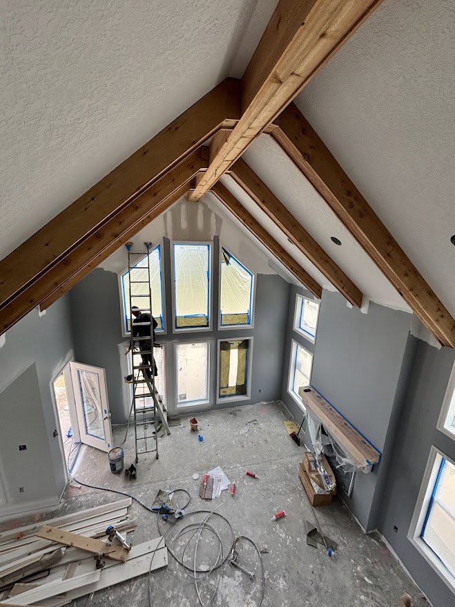 Man on ladder installing wooden beam in unfinished room with plaster walls, pile of wood planks and screwdriver on floor, white door with curtain, exposed cable, close-up of white