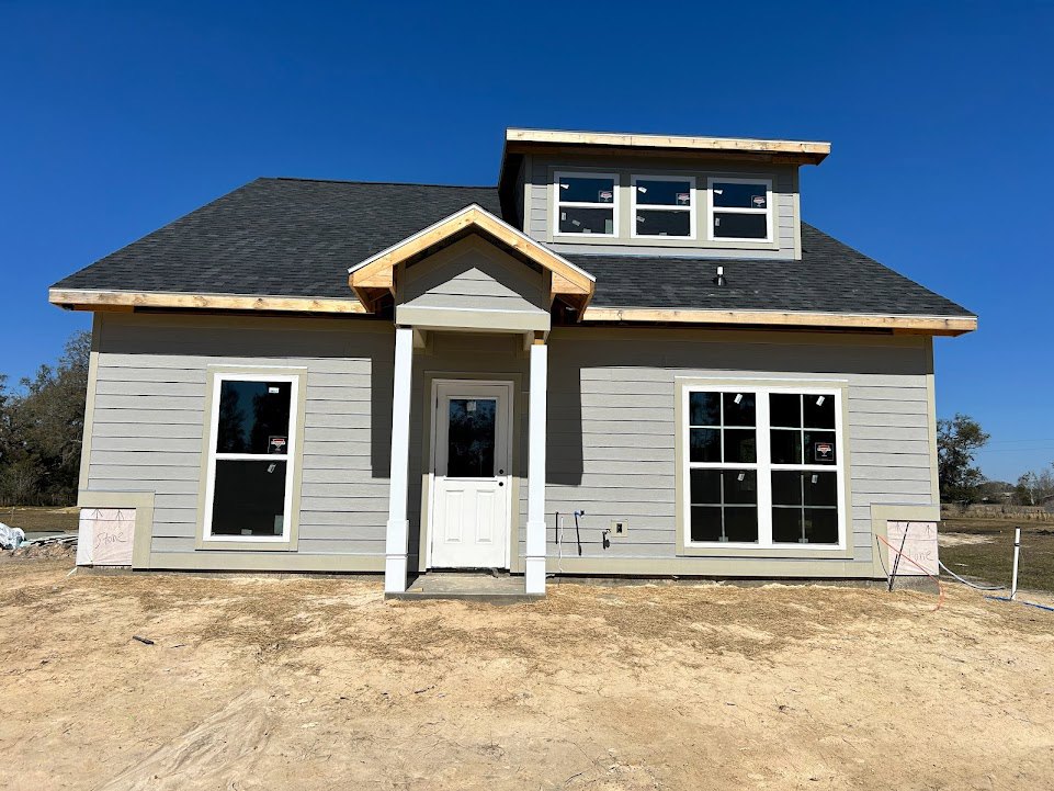 Partially built house with exposed wooden framing, white framed windows, white door with glass insert, unfinished siding, and dirt patch in front under clear blue sky