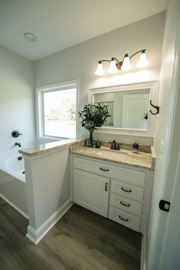 Modern bathroom featuring a freestanding bathtub, white ceramic sink on a stone countertop, tiled walls and floor, wooden cabinetry, chrome faucet, and a vase of fresh flowers.