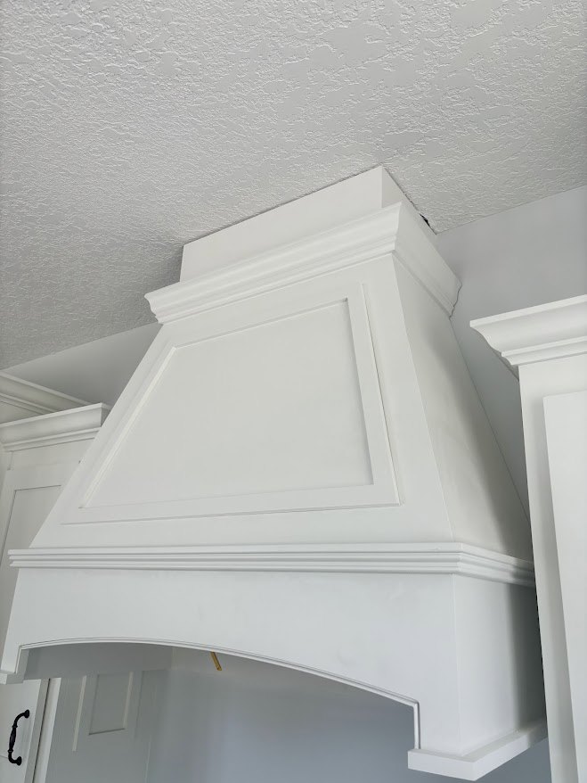 White plaster vent hood with decorative molding against a white wall and ceiling, adjacent to a white door frame in a kitchen.