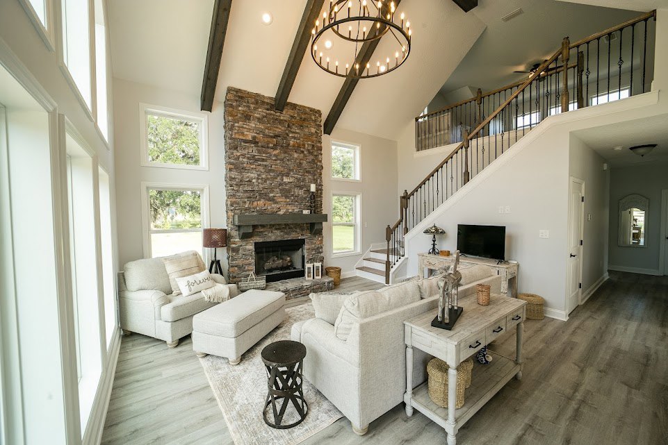 Living room with stone fireplace and wooden mantle, white couch with pillow, round coffee table, metal stool, chandelier with multiple lights, hardwood flooring