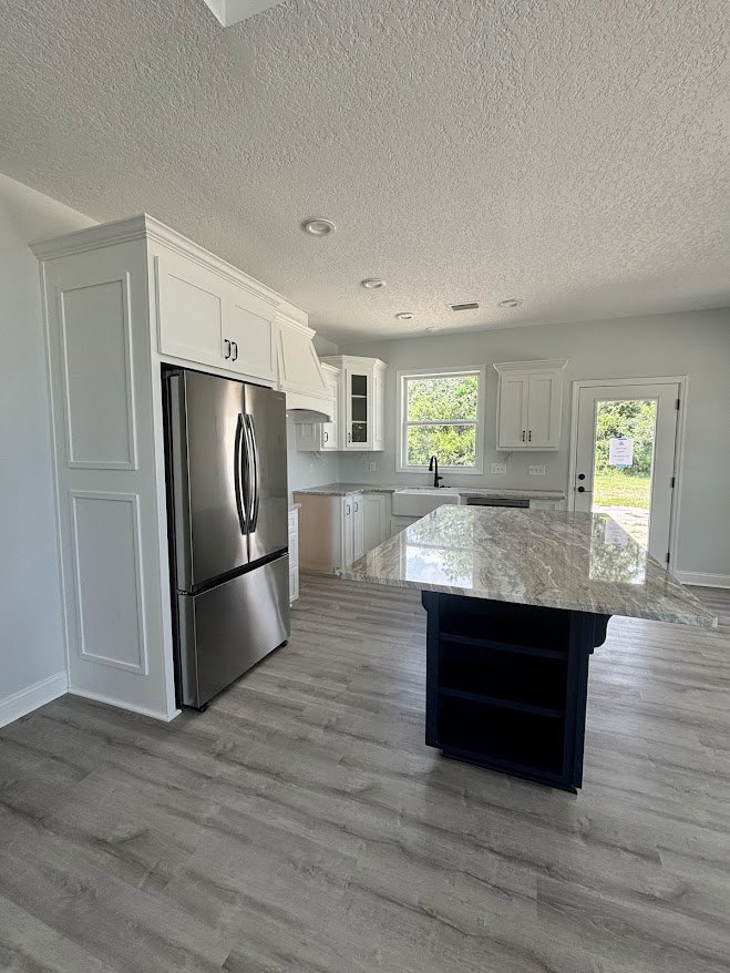 Marble island with black base, stainless steel refrigerator, white cabinetry, laminate flooring, large window overlooking trees