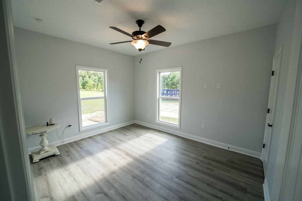 Wood flooring room with plaster walls, ceiling fan with light, white table topped with vase, large windows overlooking grassy field and blue container.