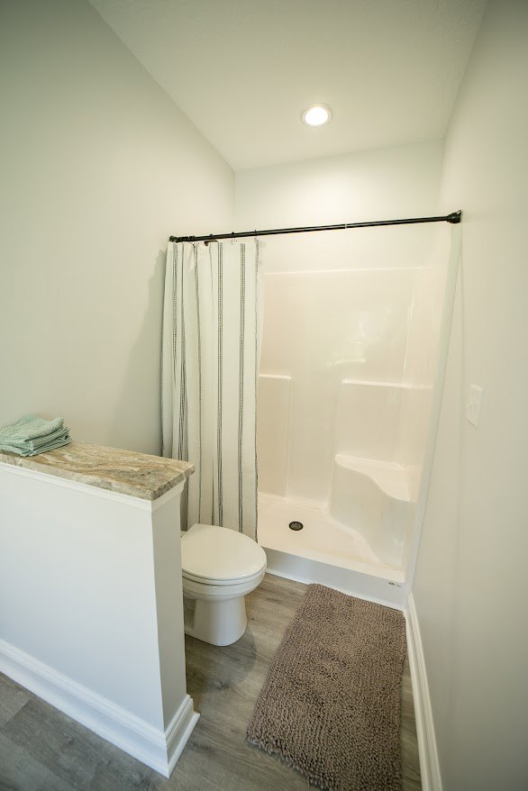 White tiled bathroom featuring a glass-enclosed shower, closed white toilet against a white wall, stack of folded towels on a brown carpet over wood flooring, and a close-up of a