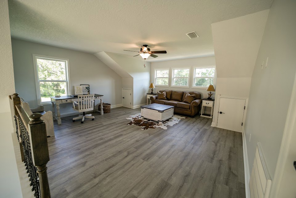 Brown couch with patterned pillows beside a wooden coffee table, desk and cushioned chair near white door with black handle, laminate wood flooring, ceiling fan with light fixture