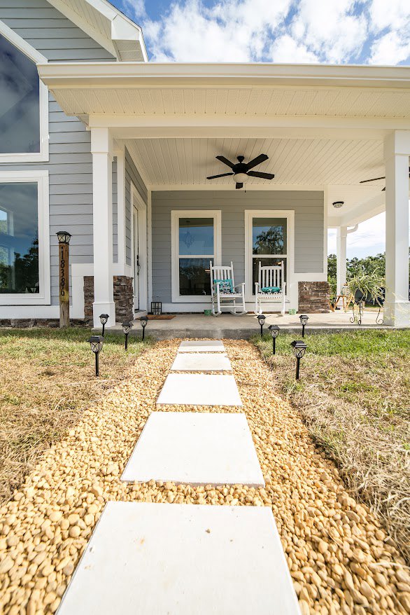 Stone walkway leading to a landscaped lawn and porch, white pillars with trim, outdoor seating featuring a white chair with a blue pillow, ceiling fan with light fixture, large