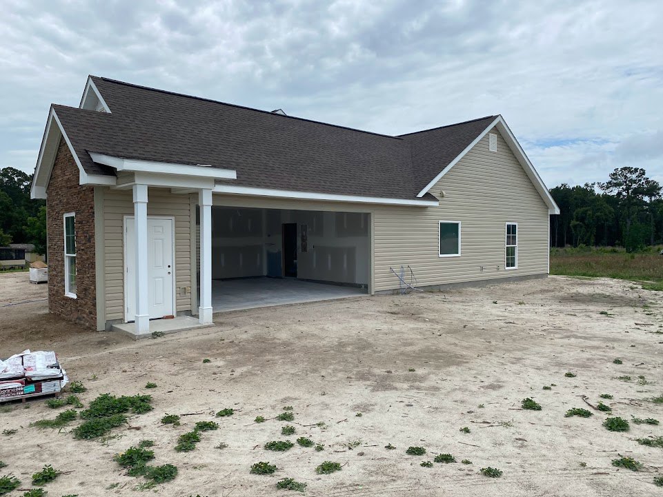Two-story custom home with attached garage, light-colored siding, shingle roof, paved driveway, landscaped front yard with shrubs and young trees under partly cloudy sky