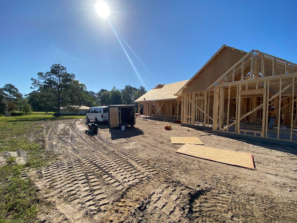 Framed house under construction with exposed wood, truck and trailer parked on dirt driveway, tire tracks visible, mature tree in front yard, roof partially finished, blue sky