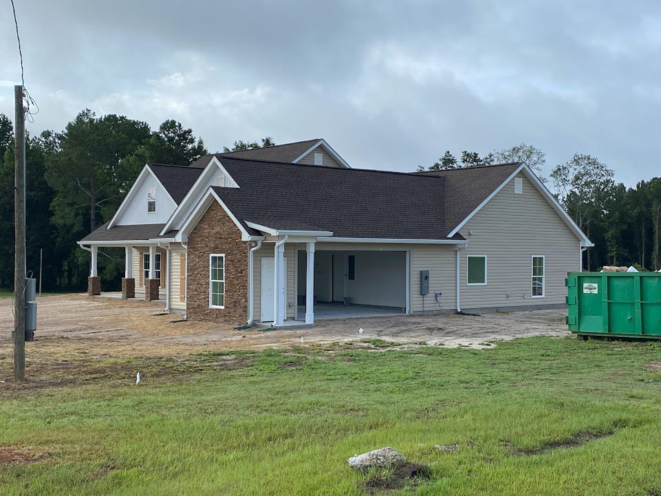 Partially built house with exposed framing, attached garage, green lawn in foreground, green dumpster on trailer, construction materials scattered, utility pole with sign, cloudy