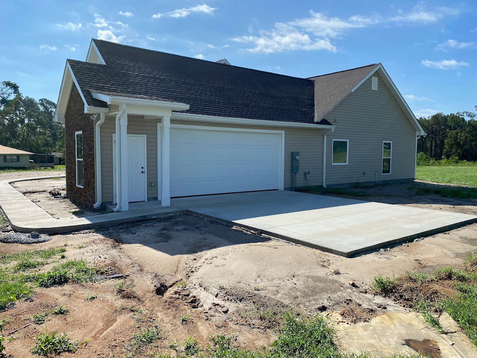Partially built house with exposed framing, attached garage featuring white door with black hardware, concrete driveway, person standing near unfinished landscaping, cloudy sky