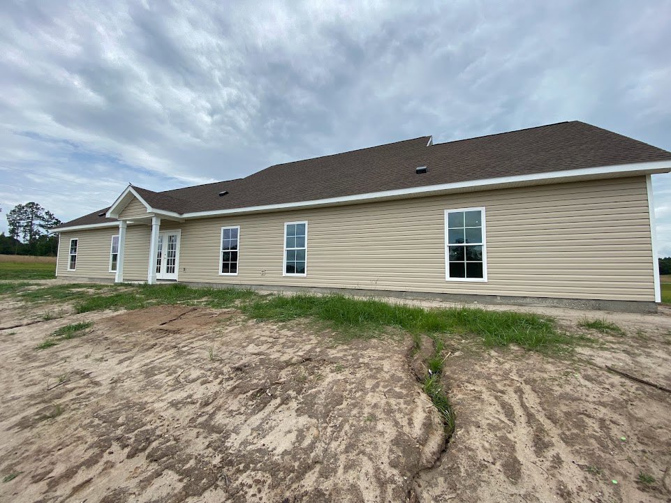 Two-story house with gray siding, white-framed windows, dark shingle roof, dirt yard with sparse grass, cloudy sky, and trees in the background
