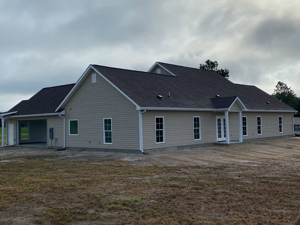 Partially built house with gray siding, shingled roof, large windows, and patchy grass under a cloudy sky, trees visible in the background