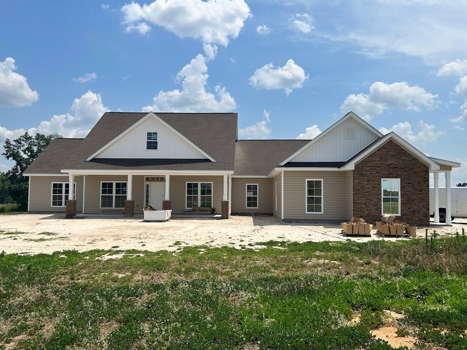 Two-story custom home with gray siding, gabled roof, covered front porch, large grassy yard, scattered cardboard boxes, and blue sky with white clouds