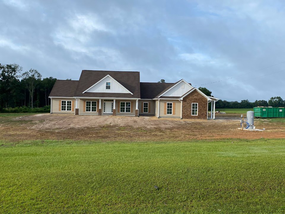 Two-story farmhouse with white siding, brick chimney, blue picket fence, manicured lawn, and surrounding trees under a cloudy sky
