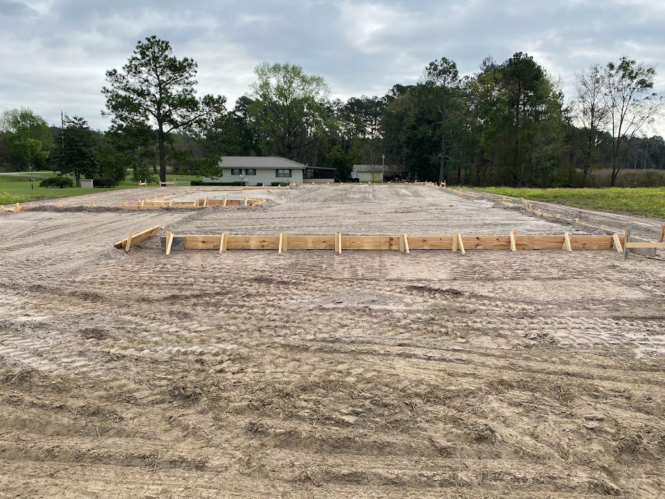 Partially built custom home framed with wooden beams on a grassy lot, surrounded by mature trees under a cloudy sky
