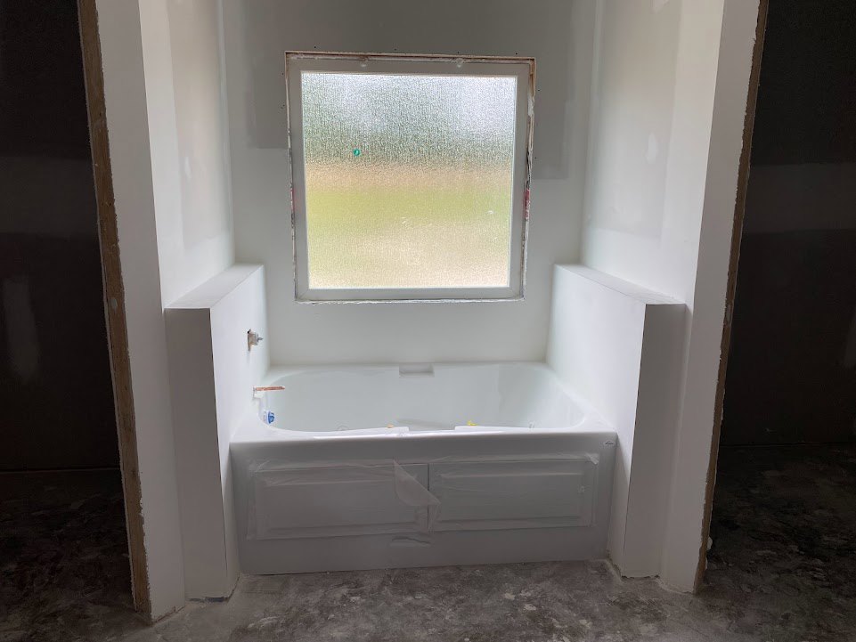 Freestanding white bathtub beside frosted glass window, light plaster walls, wood vanity with drawer, tile floor