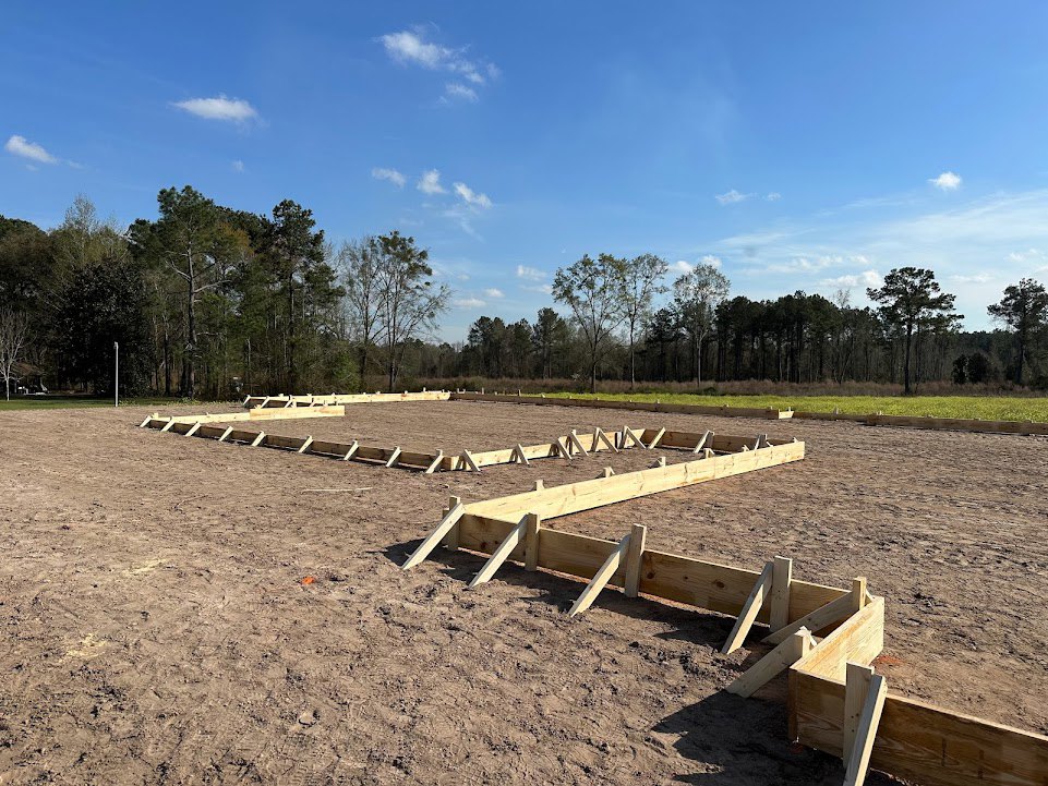 Wooden foundation frame set in dirt, surrounded by grassy field and trees under a blue sky with scattered clouds
