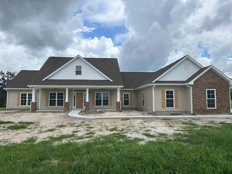 Two-story house with white roof and white-framed windows, surrounded by green lawn under a partly cloudy blue sky, car parked in background