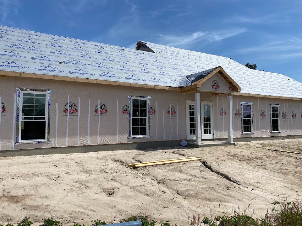 Partially built house with white door featuring glass panes, multi-pane window, exposed roof, wooden planks scattered on sandy dirt area, light-colored siding, cloudy sky overhead