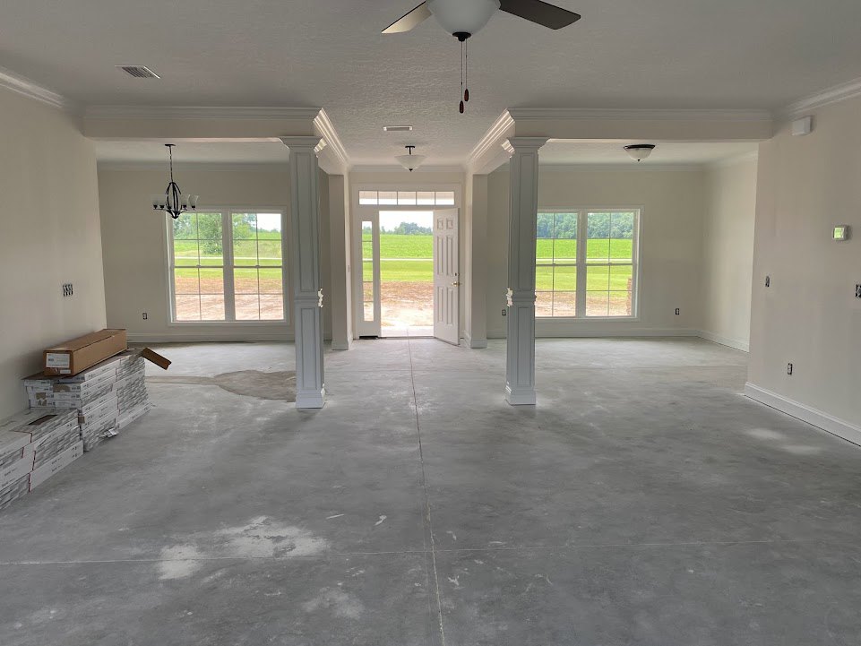 Open living space with plaster columns, ceiling fan, concrete flooring, large window overlooking green lawn, and door opening to a field